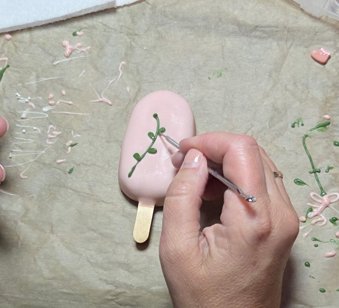 Hand painting a floral design on a pink cakesicle